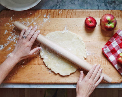 A person rolls out dough on a wooden board with a rolling pin. There are red apples and a red checkered cloth beside the board. The surface is lightly dusted with flour.