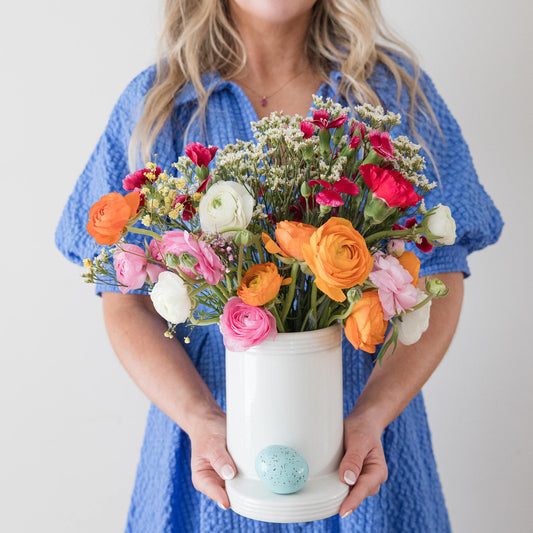 Person holding a colorful bouquet of flowers in a white vase against a plain background