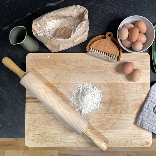 A wooden cutting board with a pile of flour and a rolling pin. Nearby are a paper bag of flour, a small brush, a carton with eggs, and a checkered cloth, all set on a dark countertop.