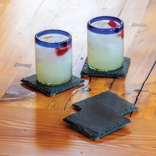 Two cocktails with blue rims on black slate coasters on a wooden surface.