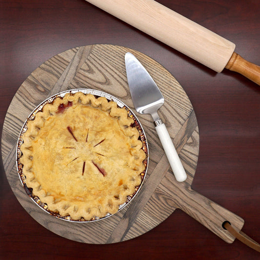 A freshly baked pie with a golden crust sits on a round wooden board. A pie server rests nearby, and a wooden rolling pin is placed at the top of the board, all on a dark wooden surface.
