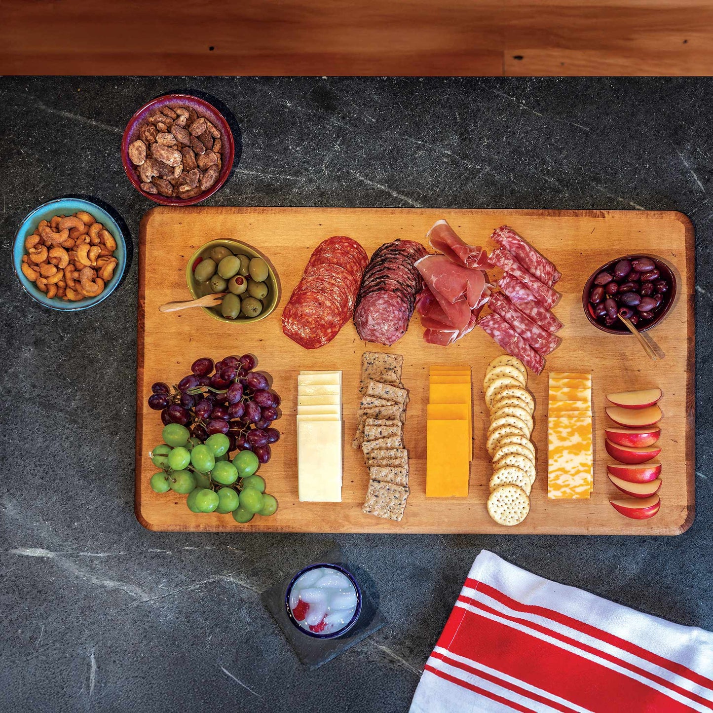 Wooden cutting board with assorted meats, cheeses, fruits, and nuts on a dark surface.