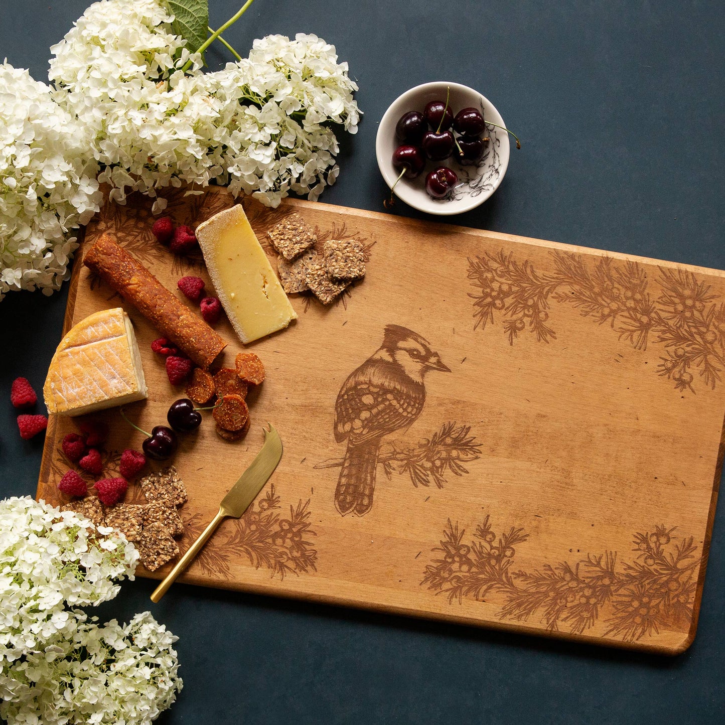 An artisan maple cheese board with an engraved blue jay. The board has cheese, crackers and charcuterie on it.