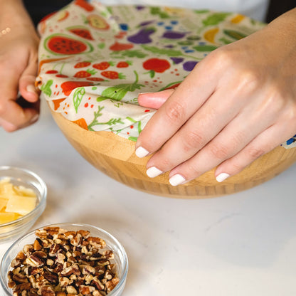 Person using a reusable food wrap on a wooden bowl with ingredients nearby.