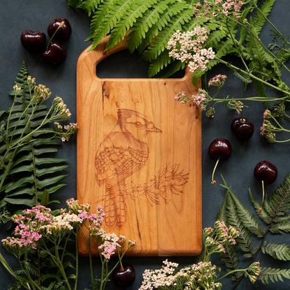 A cherry small cheese board engraved with a Blue Jay.  The board is nestled in a bed of ferns with cherries around it.
