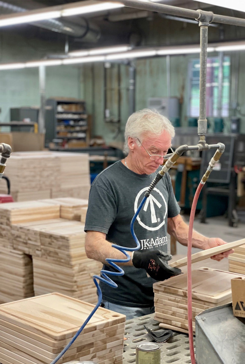 A man wearing safety glasses and a dark gray t-shirt with a white logo works in a woodworking shop, using a pneumatic tool on a stack of wooden boards.