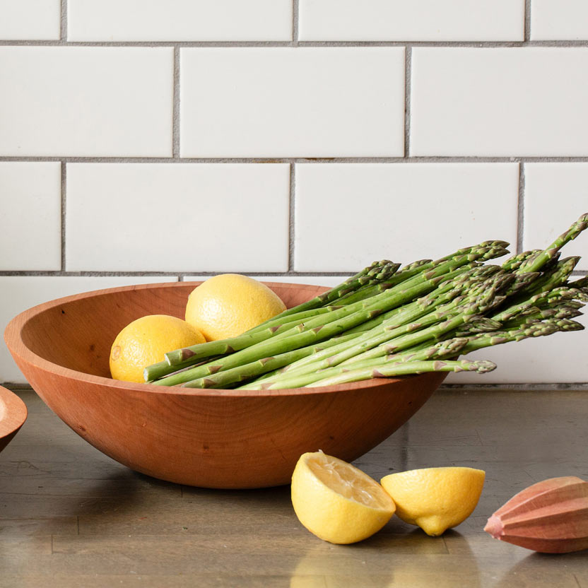 A round wooden bowl filled with green asparagus and yellow lemons, set on a countertop against a white subway tile backsplash.