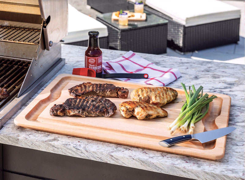 Grilled meats on a cutting board with a bottle of barbecue sauce and green onions on a patio.