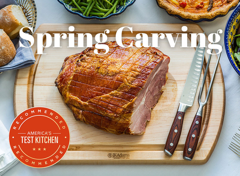 Roasted ham on a wooden cutting board with carving tools, surrounded by other food items, on a table.  Americas Test Kitchen badge.