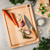 Wooden maple cutting board with carrots, shallots, garlic, and a knife on a light countertop.