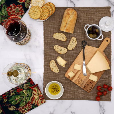 A table setting with sliced bread, cheese on a cutting board with a knife, a bowl of olives, cherry tomatoes, a glass of red wine, and a martini with olives. Crackers on a napkin with floral design complete the arrangement.
