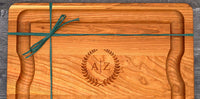 Wooden cutting board with engraved initials and laurel wreath design, tied with green twine.