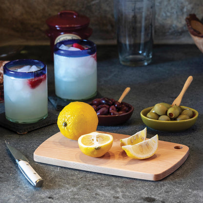 Two glasses of lemonade with ice, lemons, olives, and a knife on a wooden cutting board.