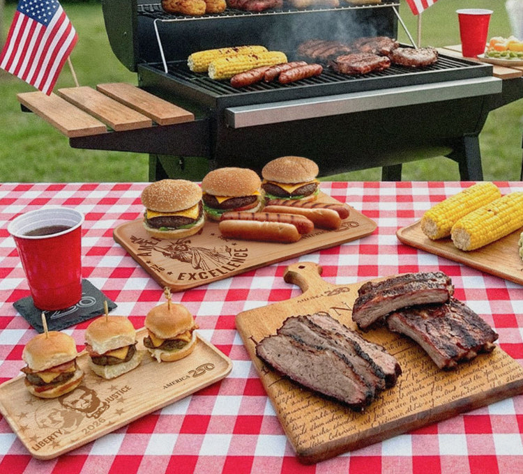 BBQ with America 250 wooden serving boards with hamburgers, ribs, and corn on the cob.
