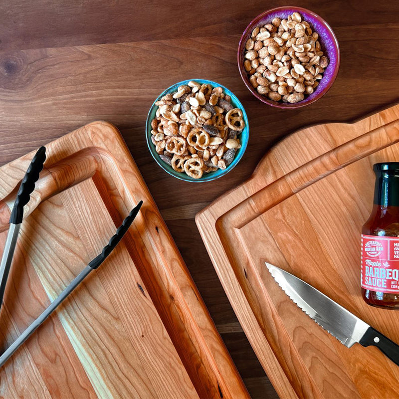 An overhead flat-lay of wooden cutting boards on a dark wood table, with a knife, a bottle of barbecue sauce, and two bowls of snacks.