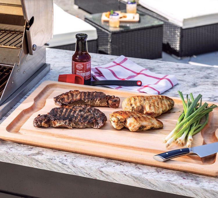 Grilled meats on a cutting board with a bottle of barbecue sauce and green onions on a patio.