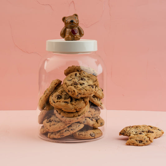 A large glass canister with a white melamine lid with a Nora Fleming teddy bear mini, filled with chocolate chip cookies. 
