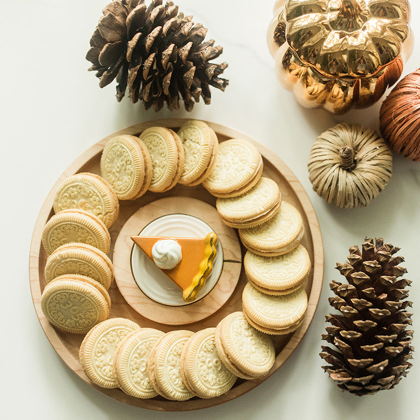 A styled flat-lay of Oreo-style sandwich cookies arranged in a circle on a round wooden board, with a small orange and white dessert slice in the center. The scene is decorated with pinecones and small decorative pumpkins on a light neutral background.