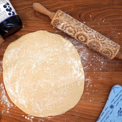 Dough rolled out in a circle on a wooden surface, imprinted with floral patterns. A decorative rolling pin with floral engravings and a jar of jam are nearby. A blue cloth is partially visible at the edge.