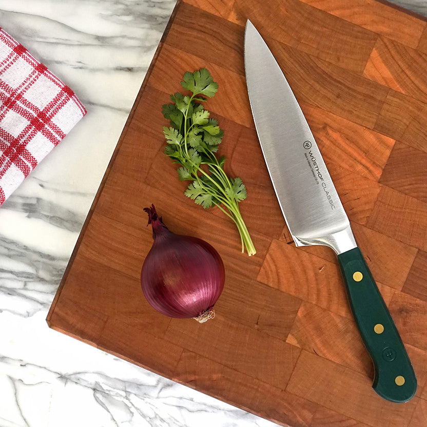 A Wusthof chef's knife with a green handle and gold dots, resting on a wooden cutting board next to a red onion and cilantro, with a red and white checkered kitchen towel on a marble surface.