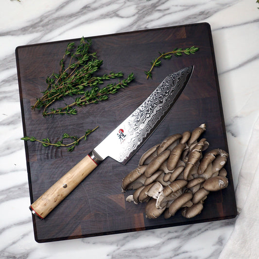 JK Adams Professional Walnut 12"x12" cutting board with herbs and mushrooms on a marble countertop.