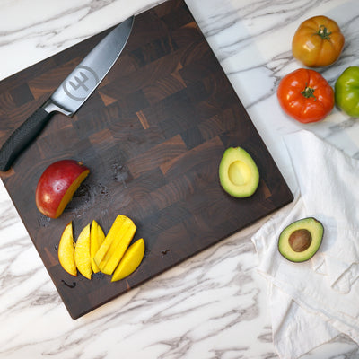 A knife and mango slices are on a wooden cutting board. A half avocado is beside a red mango, and another half avocado is on a cloth. Three tomatoes—orange, red, and green—are on the marble countertop.