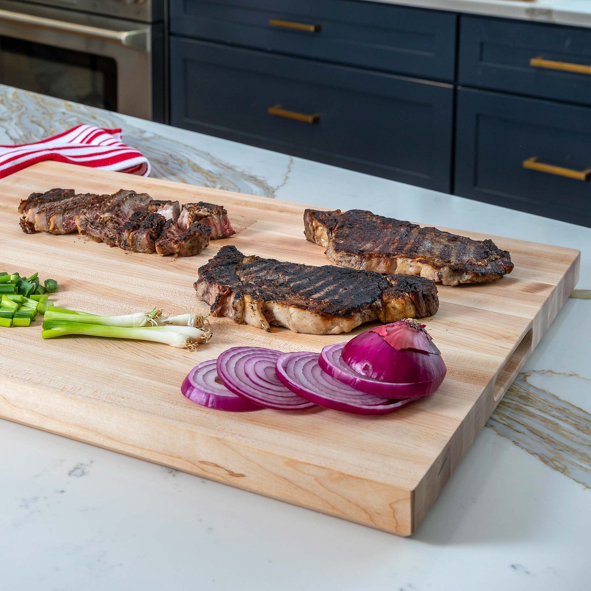 A large rectangular wooden cutting board with a built-in handle, shown on a kitchen counter with grilled steaks, sliced red onion, green onions, and a striped cloth.