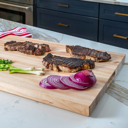 A large rectangular wooden cutting board with a built-in handle, shown on a kitchen counter with grilled steaks, sliced red onion, green onions, and a striped cloth.