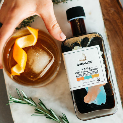 A HAND REACHING FOR AN OLD FASHIONED IN A GLASS NEXT TO A BOTTLE OF OLD FASHIONED COCKTAIL MIXER