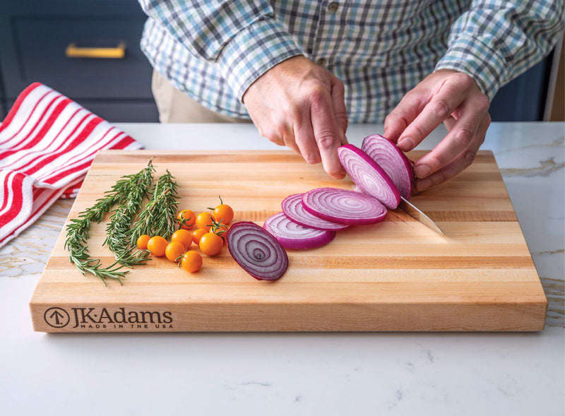 Person slicing red onions on a wooden cutting board with JK Adams branding.